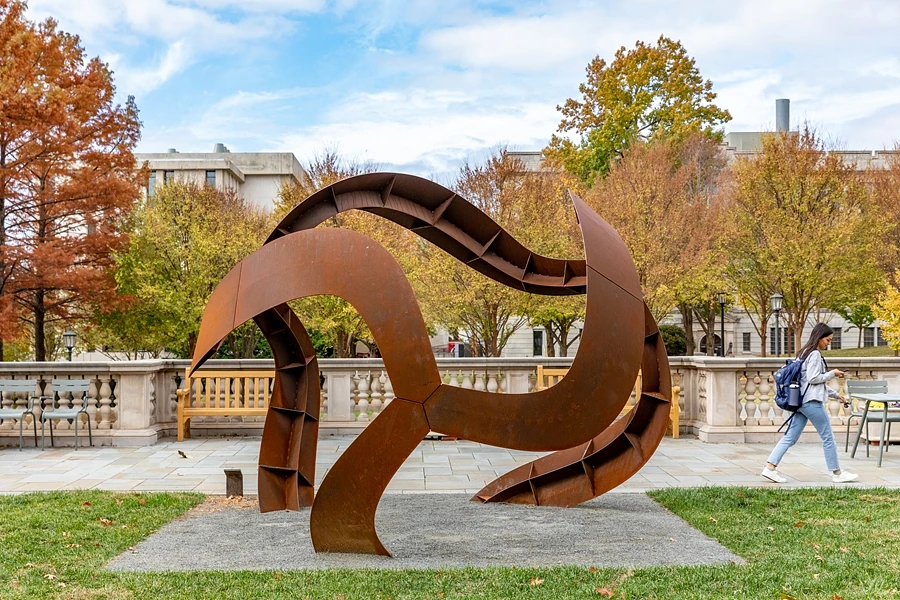 A large, abstract, rust-colored metal sculpture is displayed outdoors with autumn trees in the background and a person walking nearby.